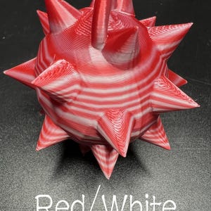 May include: A spherical object with multiple pointed spikes, featuring a layered red and white striped pattern. The surface shows visible horizontal ridges from the manufacturing process. The text Red/White is printed below the object.