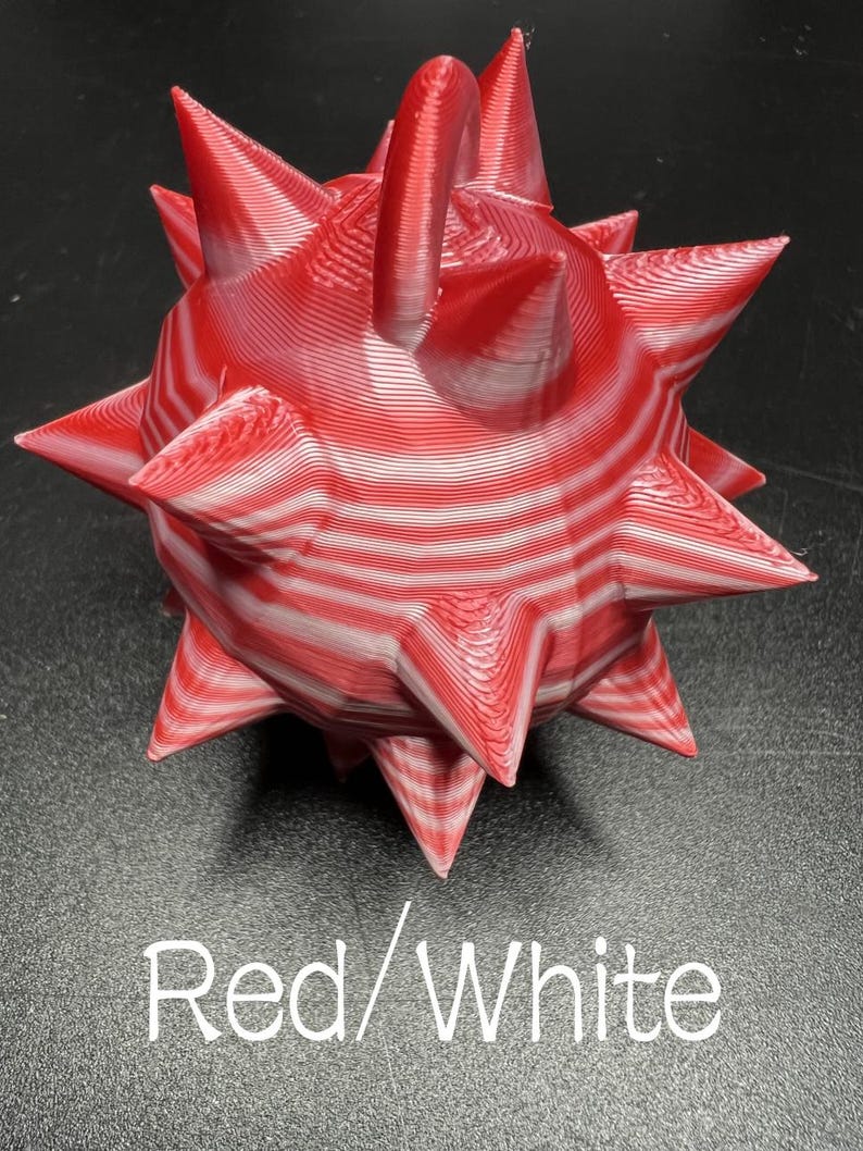 May include: A spherical object with multiple pointed spikes, featuring a layered red and white striped pattern. The surface shows visible horizontal ridges from the manufacturing process. The text Red/White is printed below the object.