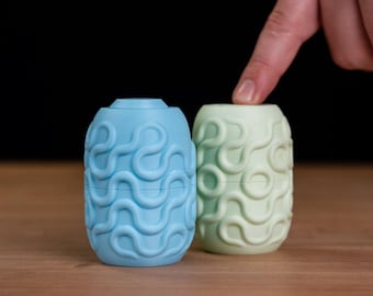 May include: Two cylindrical containers with a matte finish, one light blue and one pale green, both featuring a raised, continuous swirling line pattern that wraps around the exterior surface of each object.