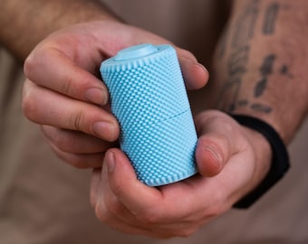 May include: A cylindrical light blue object with a textured, studded surface. The item features a flat top with a circular center indentation and a horizontal seam dividing the body into two sections, creating a tactile, grip-friendly exterior.