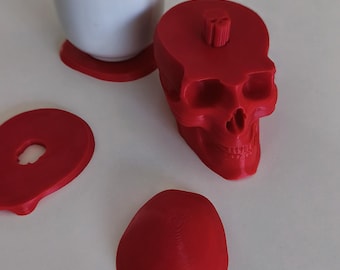 May include: A bright red 3D-printed skull-shaped container with a removable lid, featuring a small knob on top. Beside it lies a separate flat red circular piece with a central hole, and a white ceramic cup resting on a red coaster.