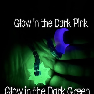 May include: A glow in the dark pink ring and a glow in the dark green spiked ball pendant on a chain. The text Glow in the Dark Pink appears at the top and Glow in the Dark Green appears at the bottom of the image against a black background.