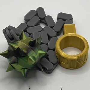 May include: A 3D printed set featuring a dark grey chain, a green and black spiked ball, and a gold ring with a tab. The text Black/Yellow/Green is printed at the bottom. The objects have a layered texture from the printing process.