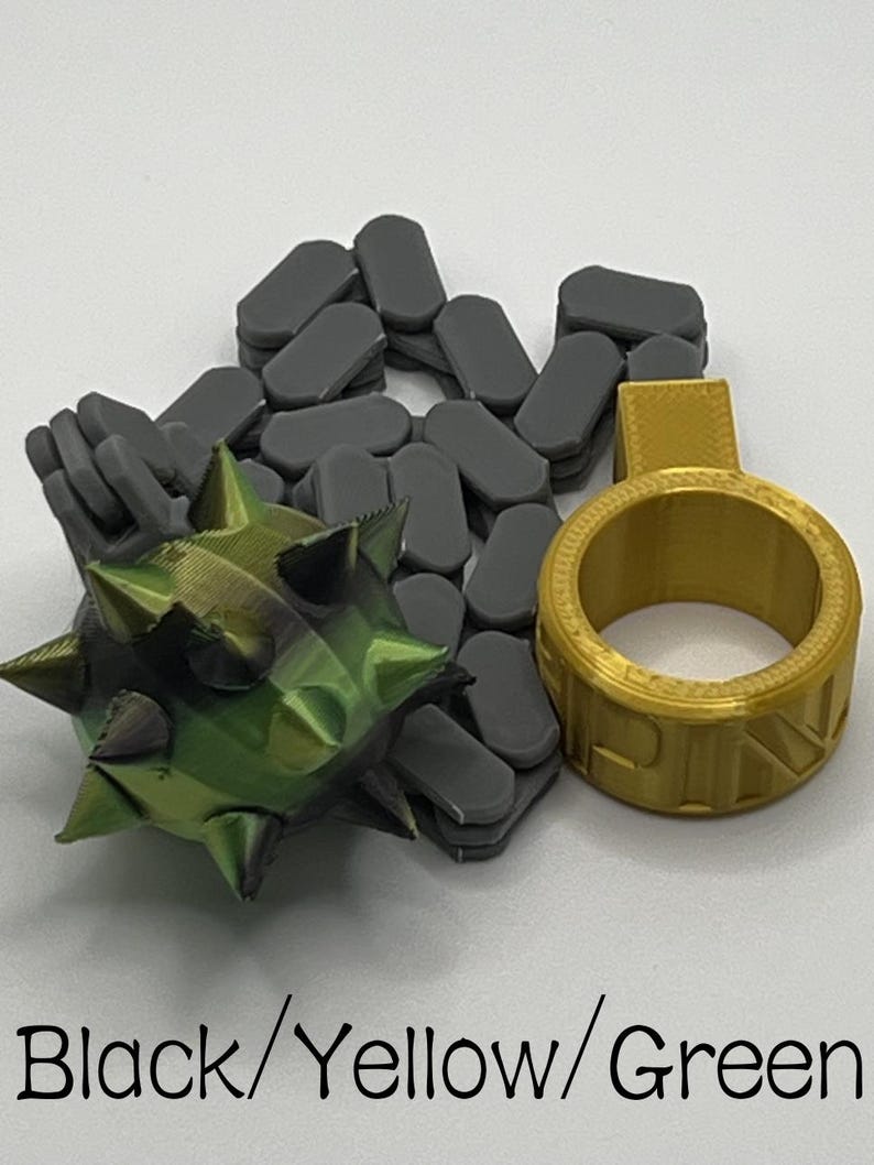 May include: A 3D printed set featuring a dark grey chain, a green and black spiked ball, and a gold ring with a tab. The text Black/Yellow/Green is printed at the bottom. The objects have a layered texture from the printing process.