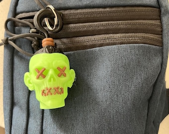 May include: A dark blue canvas sling bag with multiple zippered compartments and an M-TAC logo patch. Attached to the zipper pull is a neon green skull-shaped charm featuring brown stitched X marks over the eyes and mouth, secured by a ring.