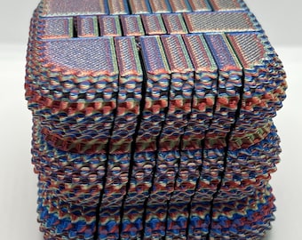 May include: A stack of square, multi-colored coasters featuring a 3D-printed, layered texture. The surface displays a repeating pattern of rectangular and square segments in a gradient of blue, red, and gold hues with visible ridges.
