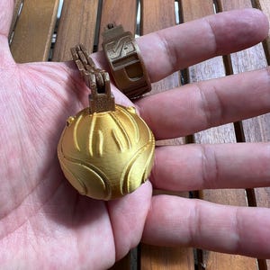 Peut inclure: Un pendentif doré imprimé en 3D avec un design sphérique et une bague assortie. Le pendentif a une surface texturée avec des lignes courbes. La bague a un design rectangulaire avec un logo.