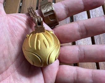 May include: A gold-colored spherical object with raised vertical ridges and a brown articulated hinge mechanism attached to a brown ring band featuring the embossed letters SD.