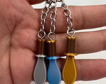 May include: Three keychains with dagger pendants in gray, blue, and gold. Each dagger has a brown handle and a silver chain. The keychains are attached to a silver keyring.