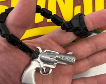 May include: A silver-colored revolver pendant with the word "Splash" engraved on the side, connected to a black chain and ring. The ring is worn on a finger. The background is a blurred image with yellow and brown colors.