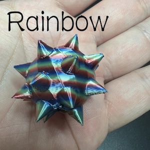 May include: A small, spherical, 3D-printed object with multiple pointed spikes, featuring a metallic rainbow gradient finish with horizontal layers. The word Rainbow is printed in black text above the object.