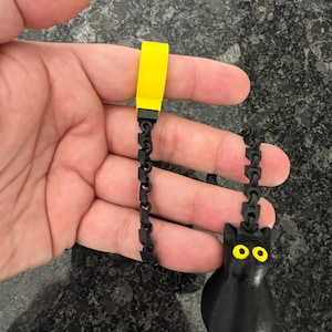 May include: A black cat-shaped pendant with bright yellow eyes attached to a black articulated chain. The chain connects to a solid yellow rectangular block at the top, creating a flexible, dangling accessory with a distinct contrast.