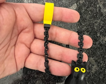 May include: A black cat-shaped pendant with bright yellow eyes attached to a black articulated chain. The chain connects to a solid yellow rectangular block at the top, creating a flexible, dangling accessory with a distinct contrast.
