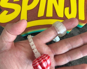May include: A translucent ring and a matching translucent chain link charm featuring a red and white striped heart-shaped pendant. The items are displayed in front of a yellow sign with the text Spinji in a bold, stylized font.