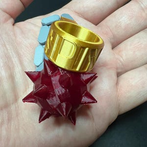 May include: A gold-coloured ring featuring a prominent, dark red, spherical mace head with multiple sharp spikes attached to the side. The band of the ring is wide and cylindrical, with the letters P A K clearly embossed on the outer surface.
