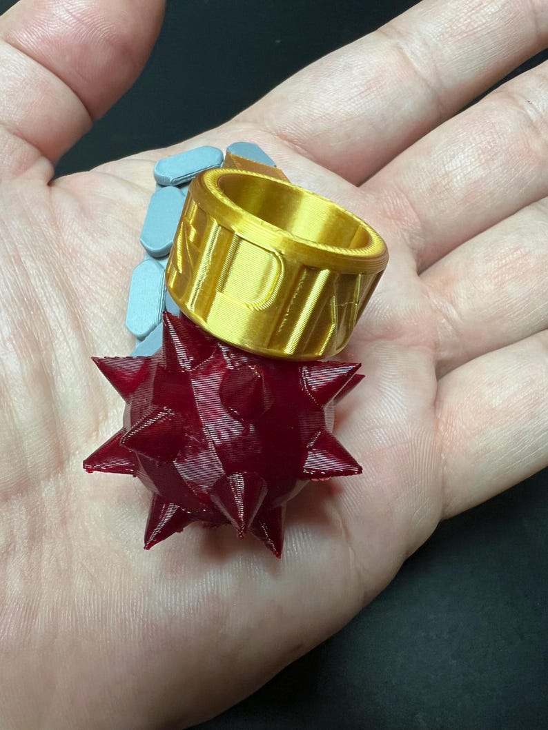 May include: A gold-coloured ring featuring a prominent, dark red, spherical mace head with multiple sharp spikes attached to the side. The band of the ring is wide and cylindrical, with the letters P A K clearly embossed on the outer surface.