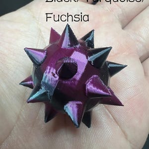 May include: A small, spherical, multi-faceted object with numerous pointed spikes. The surface features a colour-shifting gradient of black, turquoise, and fuchsia. The text "Black/Turquoise/Fuchsia" is printed above the object.