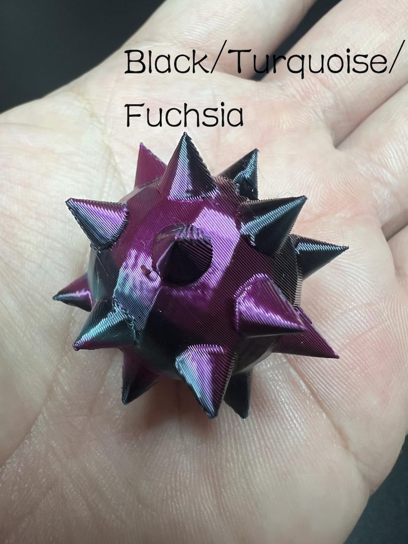 May include: A small, spherical, multi-faceted object with numerous pointed spikes. The surface features a colour-shifting gradient of black, turquoise, and fuchsia. The text "Black/Turquoise/Fuchsia" is printed above the object.