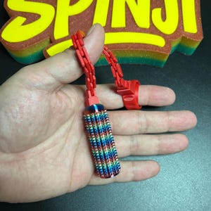 May include: A red fidget toy with a cylindrical, rainbow-coloured, spiked roller attachment. The device features a red chain and a red ring component. In the background, a large, glittery, yellow-to-green gradient sign displays the text "SPINji" in a bold, stylised font.