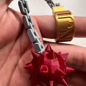 May include: A gold-coloured ring featuring a 3D-printed design with a grey articulated chain attached to a red, multi-pointed spiked ball. The ring band displays embossed lettering that appears to read 'KNY'.
