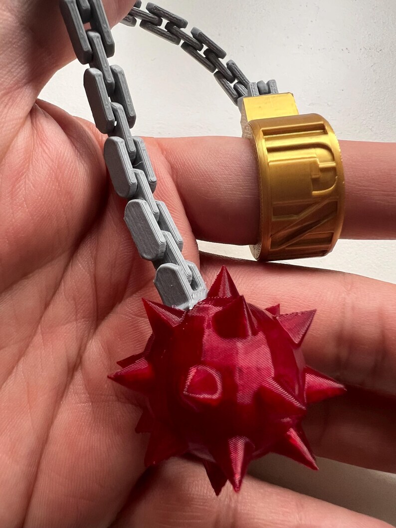 May include: A gold-coloured ring featuring a 3D-printed design with a grey articulated chain attached to a red, multi-pointed spiked ball. The ring band displays embossed lettering that appears to read 'KNY'.