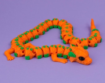 May include: An articulated dragon toy featuring a bright orange body with small green accents on each segment. The creature has a detailed head with large black eyes, small horns, and a flexible, segmented body that curves across the surface.