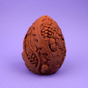 May include: A brown, egg-shaped object featuring an intricate, raised relief design of forest elements. The surface is covered in detailed carvings of oak leaves, pine cones, and acorns, creating a textured, organic appearance across the entire exterior.