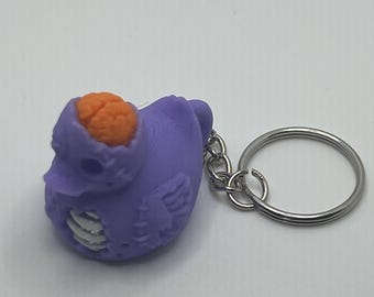 May include: A purple rubber duck keychain featuring a zombie-inspired design with an exposed orange brain on top, a hollow eye socket, and visible white rib cage detailing on the chest, attached to a standard silver-colored metal split ring.