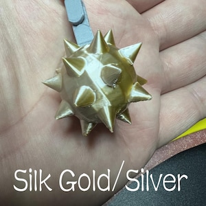 May include: A 3D printed gold-coloured spiked mace ball attached to a grey articulated chain. The text Silk Gold/Silver is printed in white at the bottom of the image. The object features a spherical shape with multiple sharp, protruding triangular spikes.