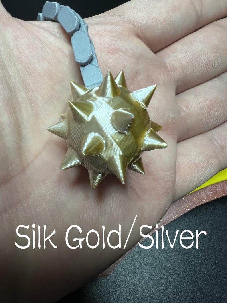 May include: A 3D printed gold-coloured spiked mace ball attached to a grey articulated chain. The text Silk Gold/Silver is printed in white at the bottom of the image. The object features a spherical shape with multiple sharp, protruding triangular spikes.