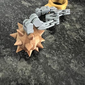 May include: A 3D printed flail toy featuring a gold-coloured spiked ball connected by a grey articulated plastic chain to a matching gold-coloured ring. The object is constructed with visible layer lines from the printing process.