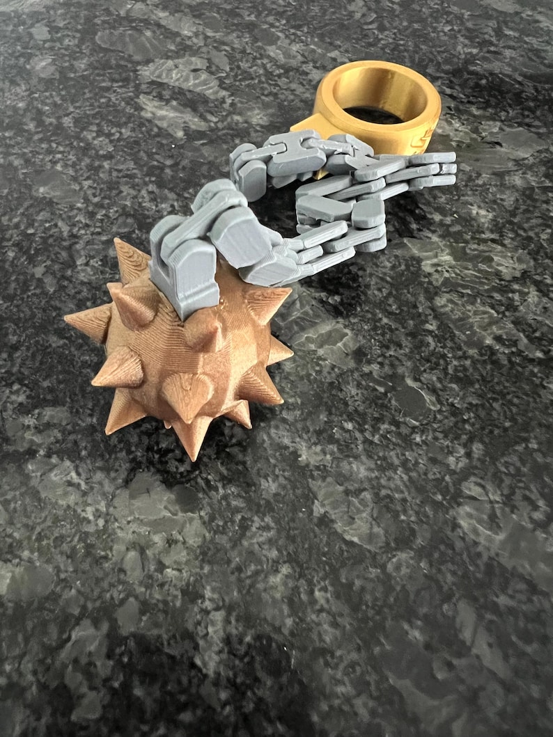 May include: A 3D printed flail toy featuring a gold-coloured spiked ball connected by a grey articulated plastic chain to a matching gold-coloured ring. The object is constructed with visible layer lines from the printing process.
