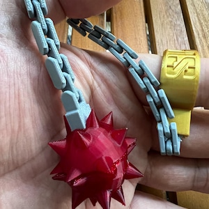 May include: A 3D printed flail toy featuring a red spiked ball attached to a grey articulated chain. The chain connects to a gold-coloured ring component that features the letters "IS" embossed on its surface. The object is constructed from plastic with visible layer lines from the printing process.
