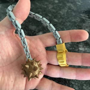 May include: A flail-style accessory featuring a gold-coloured ring connected to a grey articulated chain that ends in a bronze-coloured spiked ball. The object is constructed with a visible layered texture, indicating a 3D-printed design.