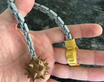 May include: A flail-style accessory featuring a gold-colored ring connected to a grey articulated chain that ends in a bronze-colored spiked ball. The object is constructed with a visible layered texture, indicating a 3D-printed design.