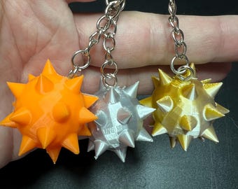 May include: Three spiked ball keychains in bright orange, metallic silver, and metallic gold. Each spherical charm features multiple pointed protrusions and is attached to a silver-toned metal chain and a circular split ring for keys.