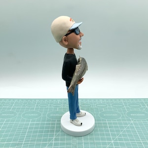 Puede incluir: Figura de un hombre con gorra beige, gafas de sol, camisa negra y vaqueros azules, sosteniendo un pez. La figura se encuentra sobre una base circular blanca. El hombre tiene barba blanca y sonr&iacute;e. El pez es gris.