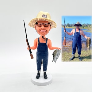 May include: A custom bobblehead figurine of a person wearing a straw hat, overalls, and holding a fishing rod and two fish. The figurine is standing on a white base.
