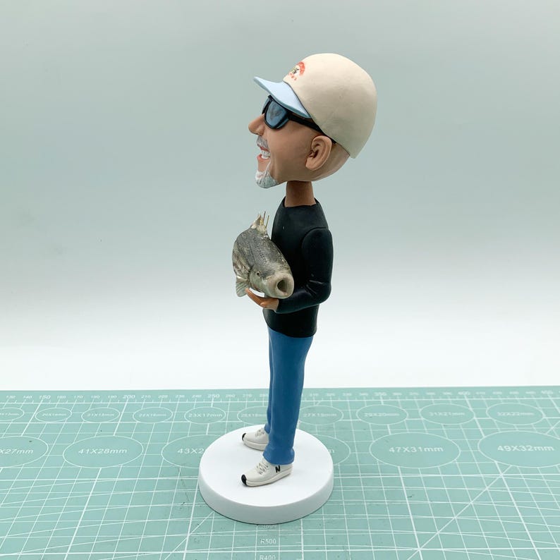 Puede incluir: Figura de bobblehead de un hombre con gorra de b&eacute;isbol y gafas de sol, sosteniendo un pez. La figura est&aacute; sobre una base circular blanca. El hombre viste una camisa negra de manga larga, vaqueros azules y zapatillas blancas.