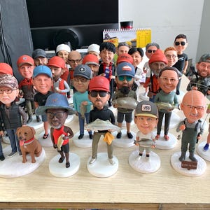 Puede incluir: Colecci&oacute;n de figuras de cabezas oscilantes, cada una representando a una persona con rasgos y vestimenta &uacute;nicos. Muchos sostienen peces, mientras que otros usan sombreros y gafas de sol. Las figuras est&aacute;n sobre bases blancas, creando una exhibici&oacute;n diversa.