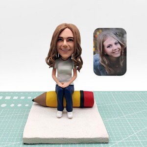 Könnte beinhalten: Eine individuelle Wackelkopf-Figur einer Person mit braunen Haaren, die ein weißes T-Shirt, blaue Jeans und weiße Turnschuhe trägt und auf einem großen gelben, braunen und roten Bleistift sitzt. Ein kleines Foto ist im Hintergrund.