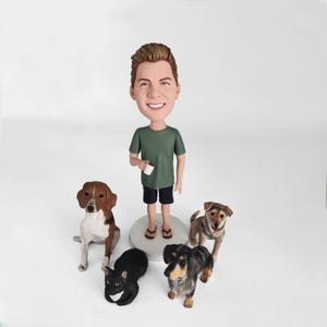 Op de afbeelding: Een gepersonaliseerd bobblehead-beeldje van een persoon met bruin haar en een groen shirt, die een witte beker vasthoudt. Het beeldje is omringd door vier kleine beeldjes van honden en een kat.