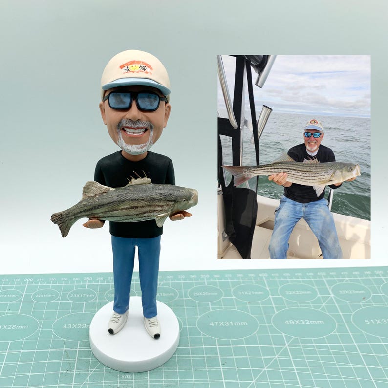 Puede incluir: Una figura de bobblehead personalizada de un hombre sosteniendo un pez rayado, con gafas de sol, un sombrero, una camisa negra, jeans azules y zapatillas blancas. Una foto m&aacute;s peque&ntilde;a muestra al hombre en un bote sosteniendo un pez similar.