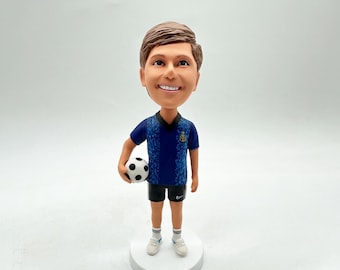 May include: A custom bobblehead figurine of a person wearing a blue and black soccer jersey, black shorts, and white socks. The person is holding a black and white soccer ball.
