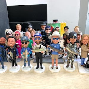 Puede incluir: Una colecci&oacute;n de figuras de cabezas oscilantes, cada una representando a una persona sosteniendo un pez. Las figuras var&iacute;an en vestimenta, con algunos usando equipo de pesca y otros ropa casual. Tambi&eacute;n hay un perro negro.