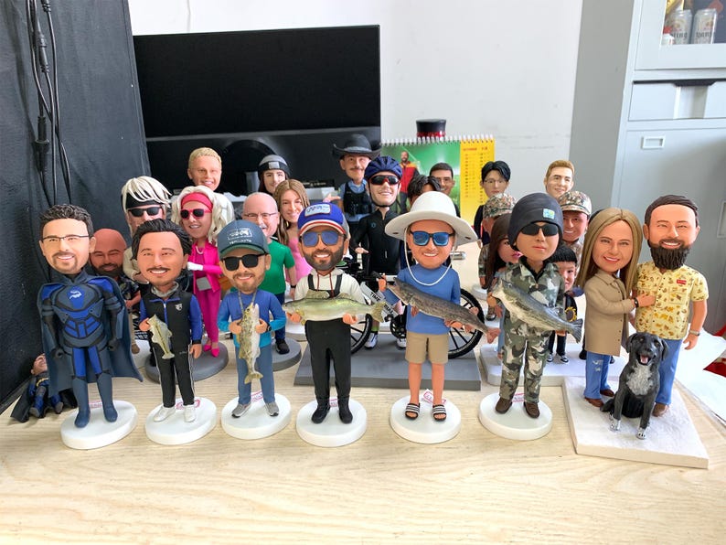 Puede incluir: Una colecci&oacute;n de figuras de cabezas oscilantes, cada una representando a una persona sosteniendo un pez. Las figuras var&iacute;an en vestimenta, con algunos usando equipo de pesca y otros ropa casual. Tambi&eacute;n hay un perro negro.
