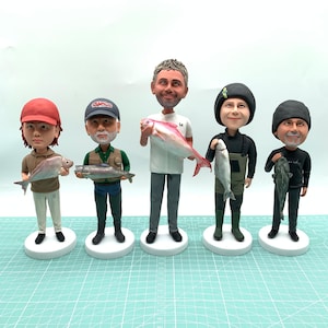 Puede incluir: Cinco figuras de cabezas oscilantes de personas sosteniendo peces. Cada figura est&aacute; sobre una base blanca y vestida con atuendo de pesca. Los peces var&iacute;an en color, incluyendo rosa y plateado. Las figuras usan sombreros, gorras y vadeadores.
