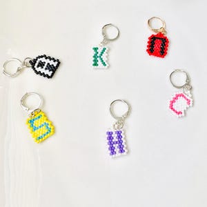 May include: Six individual charms featuring letters K, N, S, H, C, and a black and white pattern, each crafted from small colorful beads. These charms are attached to silver-toned metal hoop rings, displaying various letter designs in red, green, yellow, purple, and pink.