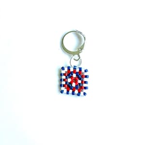 May include: A square-shaped keychain charm with a red, white, and blue pattern. The charm is made of small, square beads arranged in a grid-like design. The keychain has a silver-colored metal clasp.