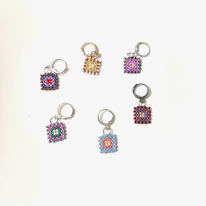 May include: A collection of square beaded earrings in various colors, including red, purple, green, and blue. Each earring features a central design and is attached to a silver-tone hoop.